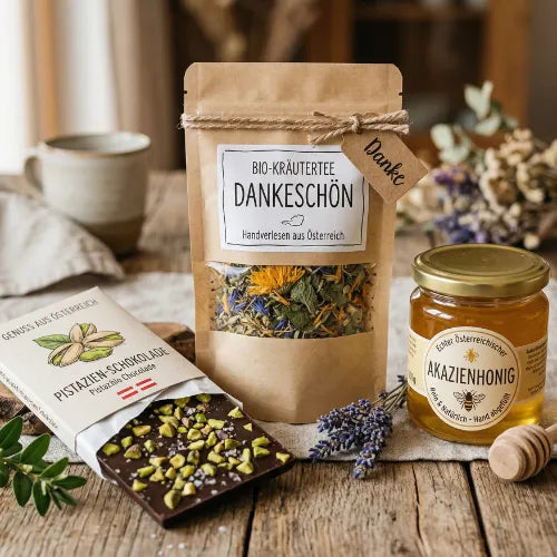 Eine Packung „Bio-Kräutertee Dankeschön“, ein Glas Akazienhonig und eine Pistazien-Schokolade auf einem Holztisch mit Lavendel.