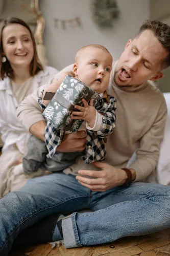 Ein lächelndes Elternpaar sitzt mit seinem Baby auf dem Boden, der Vater hält das Kind, das ein kleines, in grünliches Papier mit weißen Bäumen verpacktes Geschenk hält.
