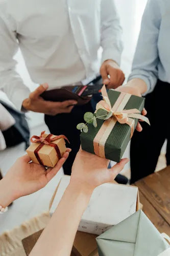 Mehrere Hände tauschen Geschenke aus. Eine Hand hält ein dunkelgrünes Paket mit cremefarbenem Band, eine andere ein kleines braunes Paket mit rotem Band.