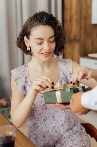 Junge Frau mit kurzen, lockigen, braunen Haaren und goldenen Ohrringen, öffnet lächelnd ein grünes Geschenk mit beigem Band an einem Holztisch.