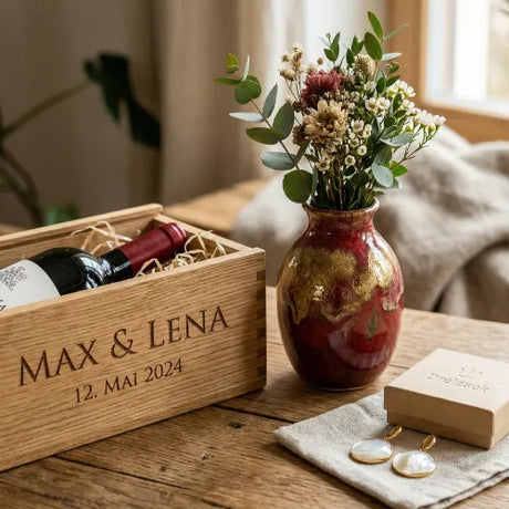 Gravierte Weinkiste mit einer Rotweinflasche, daneben eine rote Vase mit Blumen und Ohrringe in einer Schmuckschachtel auf einem Holztisch.