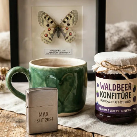 Nahaufnahme einer regionalen Waldbeer-Konfitüre, einer grünen Keramiktasse und eines silbernen, gravierten Feuerzeugs mit der Aufschrift 'MAX - SEIT 2024'.