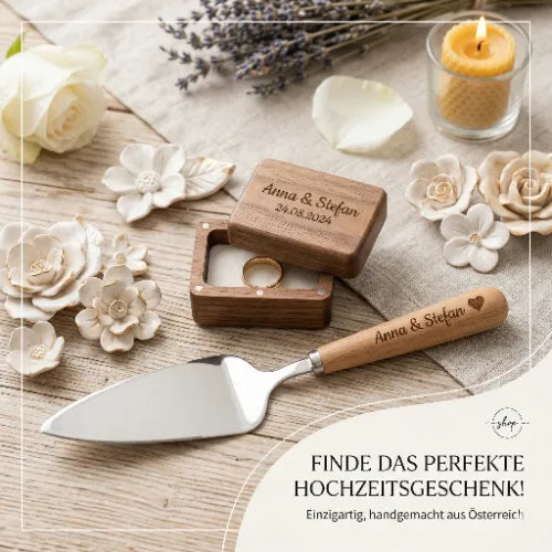Ein Ehering in einer Holzschatulle mit Gravur "Anna & Stefan 24.08.2024" und ein Tortenheber mit gleichem Namen, umgeben von weißen Keramikblüten auf hellem Holztisch