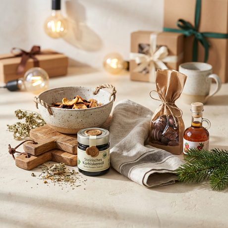Kürbiskernöl, Pralinen, Sirup, getrocknete Früchte in einer Keramikschüssel auf einem Holzbrett mit Geschenken und Tanne im Hintergrund.