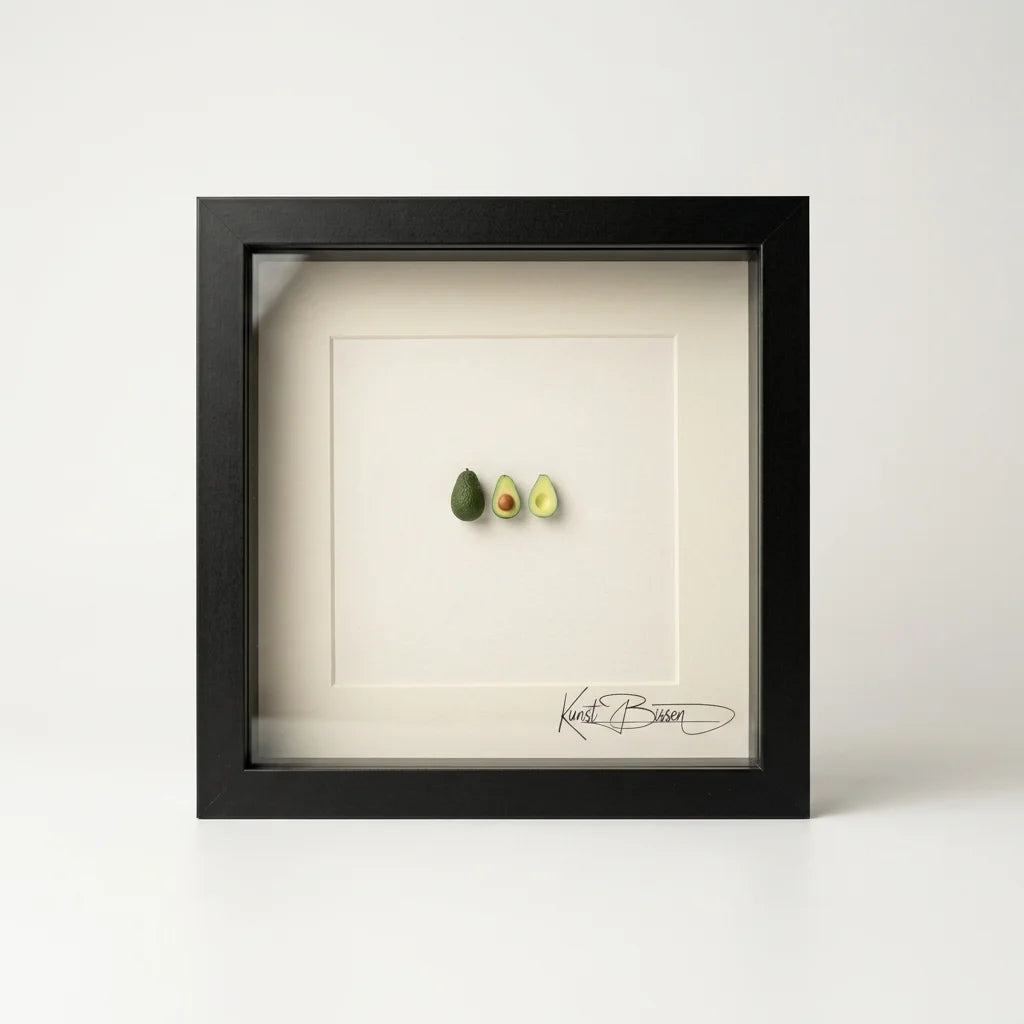 Handmodellierte Avocado-Miniatur in zwei Hälften und ganz, präsentiert in schwarzem Echtholzrahmen 22x22 cm mit Passepartout.