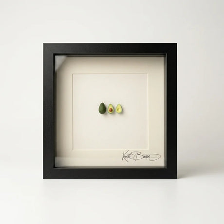 Handmodellierte Avocado-Miniatur in zwei Hälften und ganz, präsentiert in schwarzem Echtholzrahmen 22x22 cm mit Passepartout.