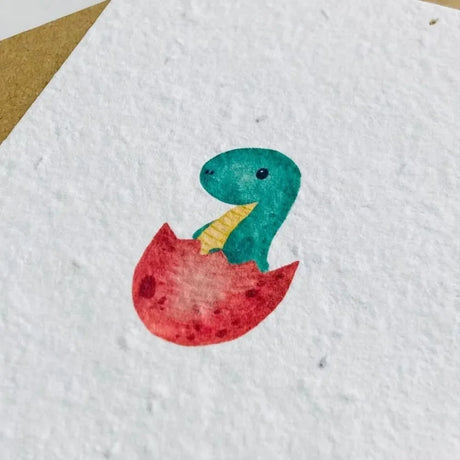 Geburtskarte aus Saatpapier mit handgemaltem Dino-Motiv, der aus einem Ei schlüpft, Schriftzug Willkommen und beigelegtem Kuvert.