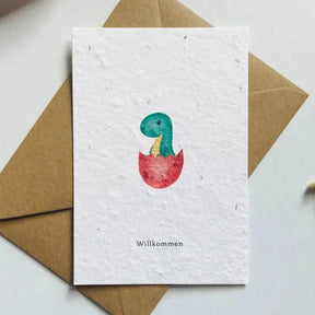 Geburtskarte aus Saatpapier mit handgemaltem Dino-Motiv, der aus einem Ei schlüpft, Schriftzug Willkommen und beigelegtem Kuvert.