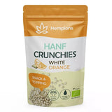 Hempions Bio Hanf Crunchies in Weiß und Orange gehalten. Detailansicht von gerösteten Hanfsamen umhüllt von weißer Kakaomasse und Orangenschalenöl in einem geöffneten Beutel. — 50g