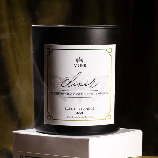 Weiße Duftkerze ELIXIR von Morii in minimalistischem Glasgefäß, umgeben von trockenen Kräutern und Blüten.