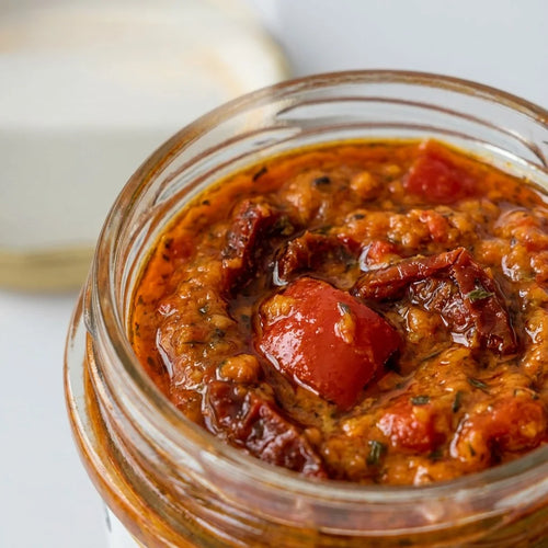 Ein Glas Paprika Knoblauch Pesto mit roter Farbe und sichtbaren Kräutern auf hellem Hintergrund.