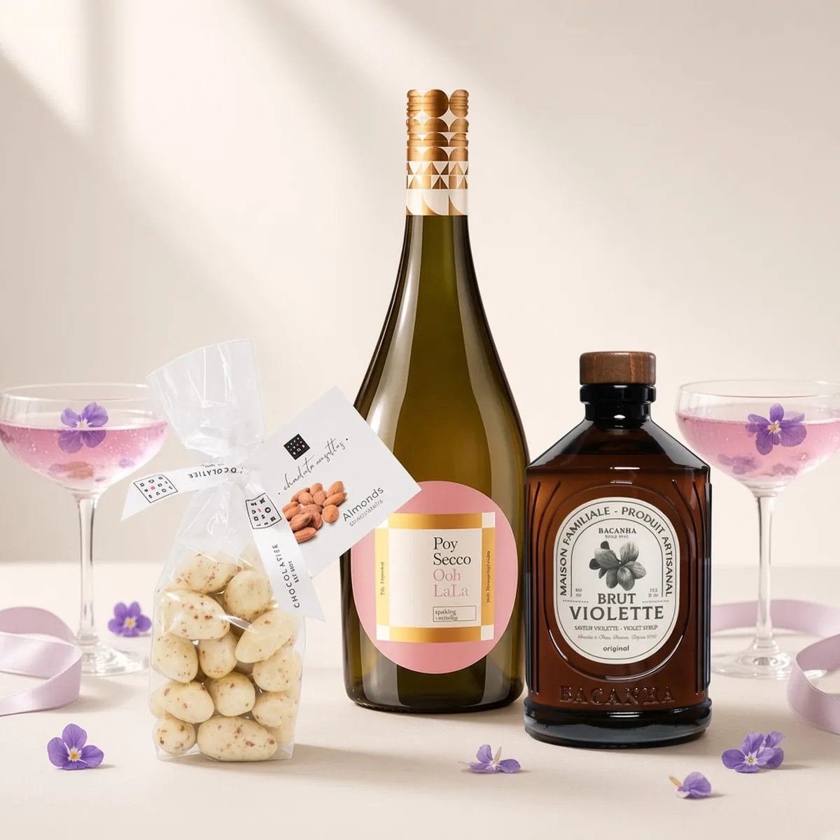 Frizzante Ooh La La mit rosa Etikett, Veilchensirup von Bacanha und Schoko-Mandeln, daneben zwei Gläser mit Veilchen