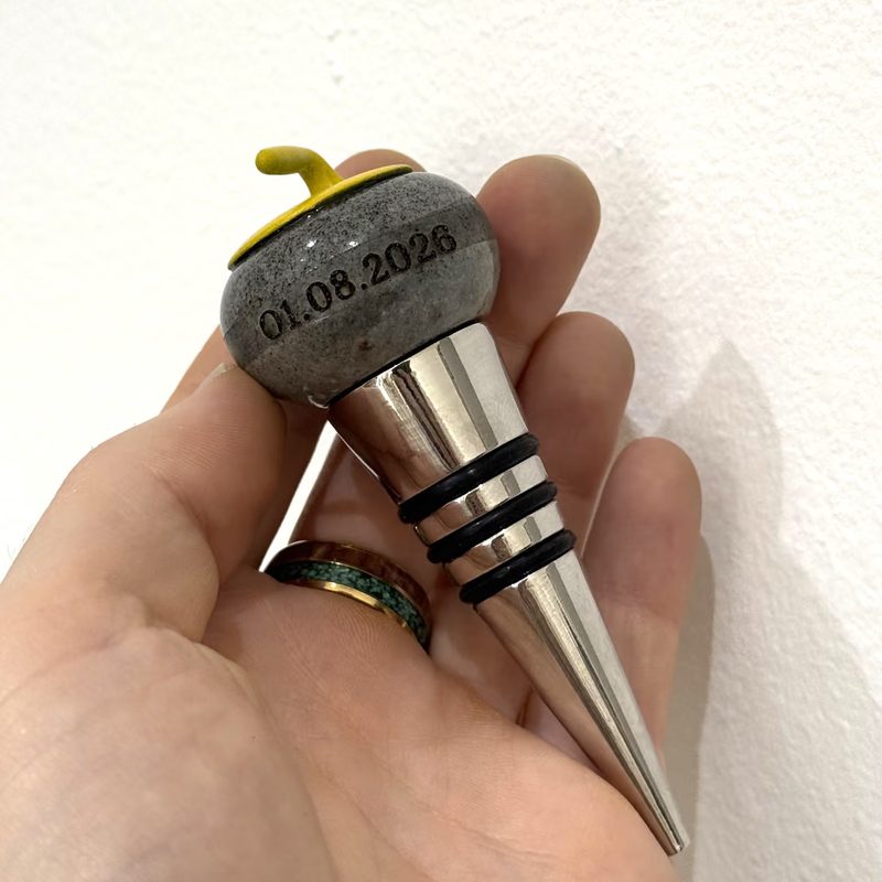 Ein Flaschenverschluss in Form eines Curlingsteins mit graviertem Datum, in der Hand gehalten