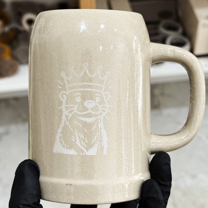 Ein Bierkrug aus heller Keramik mit eingraviertem Bär mit Krone, in behandschuhten Händen gehalten