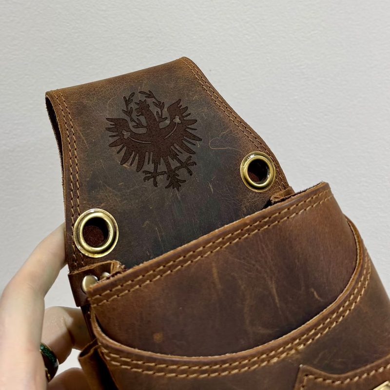 Eine braune Ledertasche mit eingraviertem Adlerwappen, in der Hand gehalten