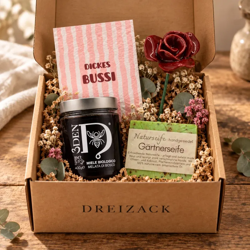 Geöffnete Dreizack Mystery Box Klein aus Kraftkarton mit Bio-Waldhonig, handgesiedeter Gärtnerseife, Keramikrose und Grußkarte „Dickes Bussi“ auf Trockenblumen
